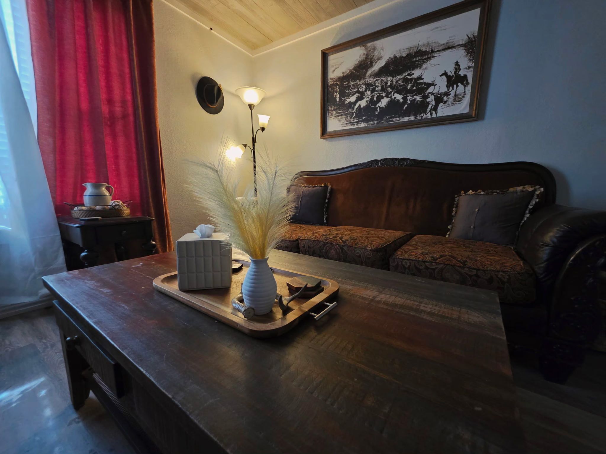 Una sala acogedora con una mesa de centro de madera, un florero con plantas secas, una lámpara y un sofá con cojines estampados.