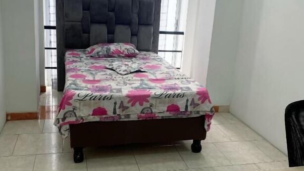 A bedroom with a bed, a window with sheer curtains, a bedside table, and a wall-mounted sign.
