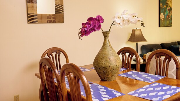 A dining table with a vase of flowers, wooden chairs, a lamp, and a wall with a mirror and artwork.