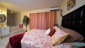 A bedroom with a bed, a nightstand, a potted plant, and a bathroom with a tub and shower.