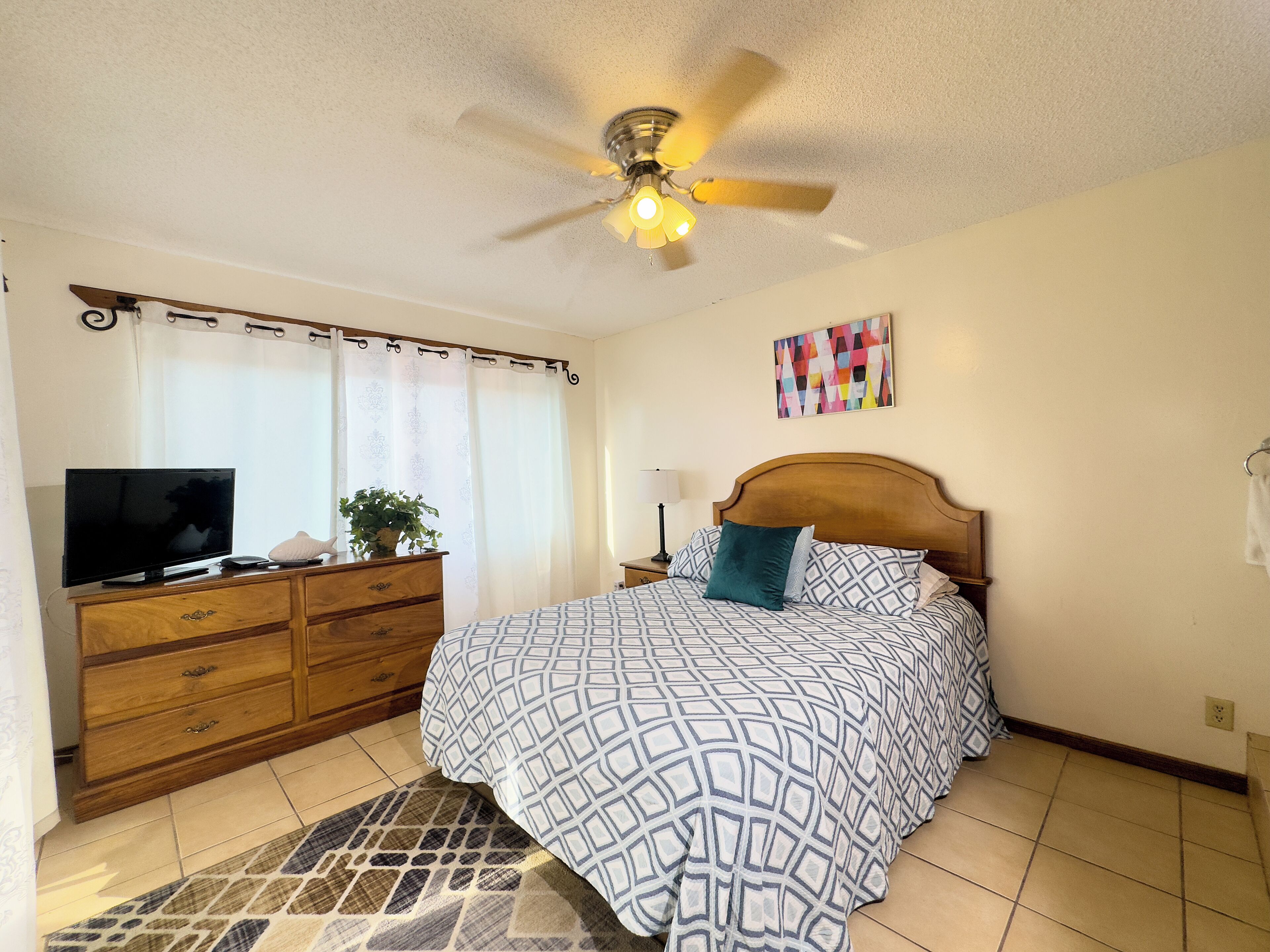 A bedroom with a bed, a TV, a dresser, a ceiling fan, and a window with curtains.
