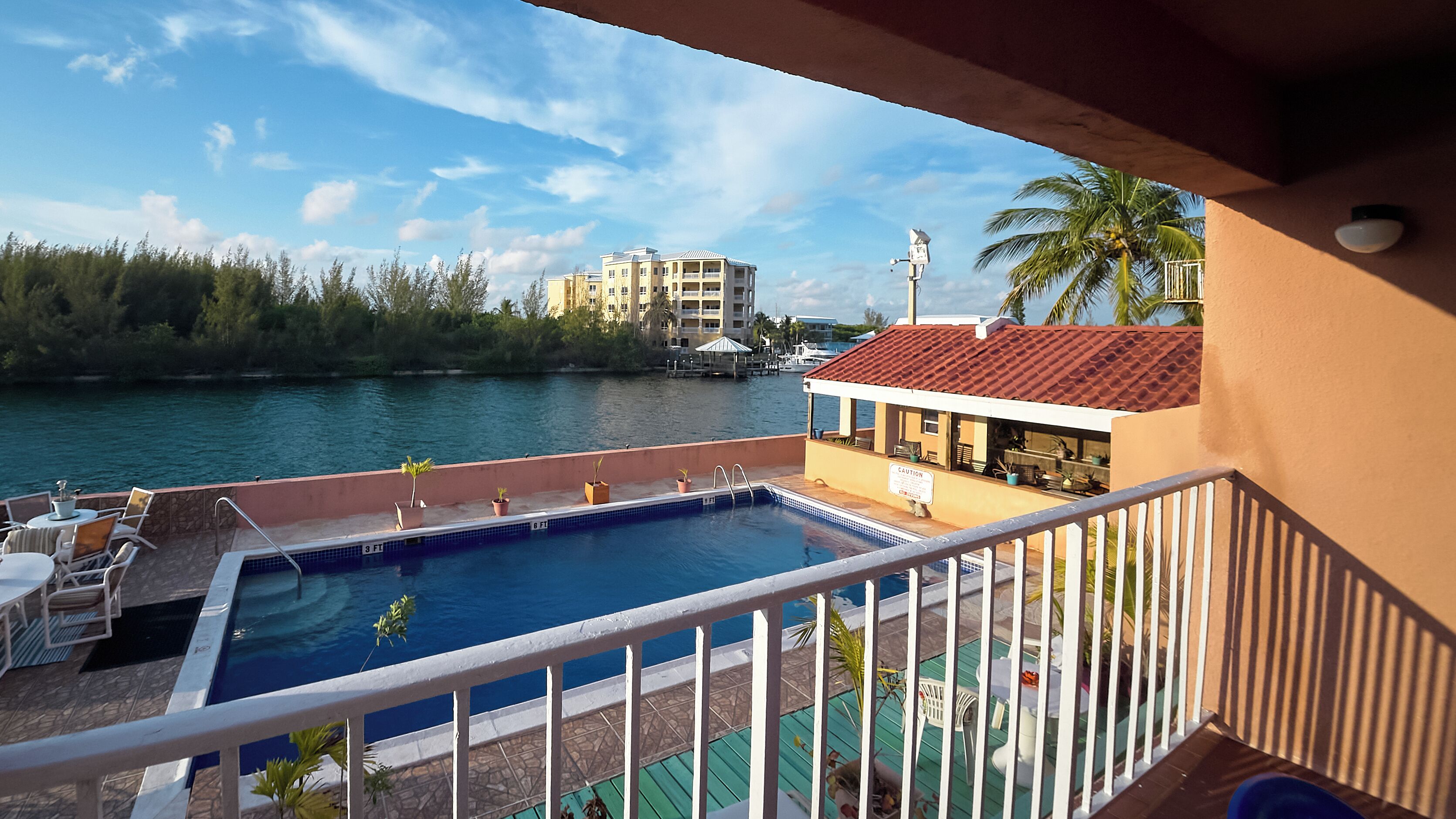 A pool area with a view of a body of water, a building with a red roof, and a palm tree.
