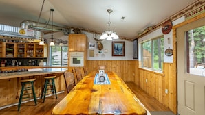 Una mesa de comedor de madera con taburetes tipo bar, una zona de cocina con refrigerador y un ventanal con vista a árboles.