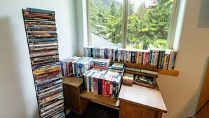 Una habitación con un ventanal grande con vistas a una zona boscosa, un escritorio de madera con libros y DVDs, y una estantería llena de diversos objetos.