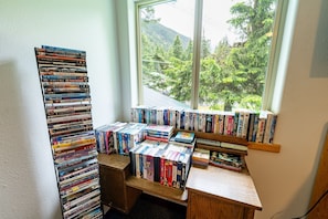 Een kamer met een groot raam met uitzicht op een bebost gebied, een houten bureau met boeken en dvd's, en een boekenplank vol met diverse spullen.