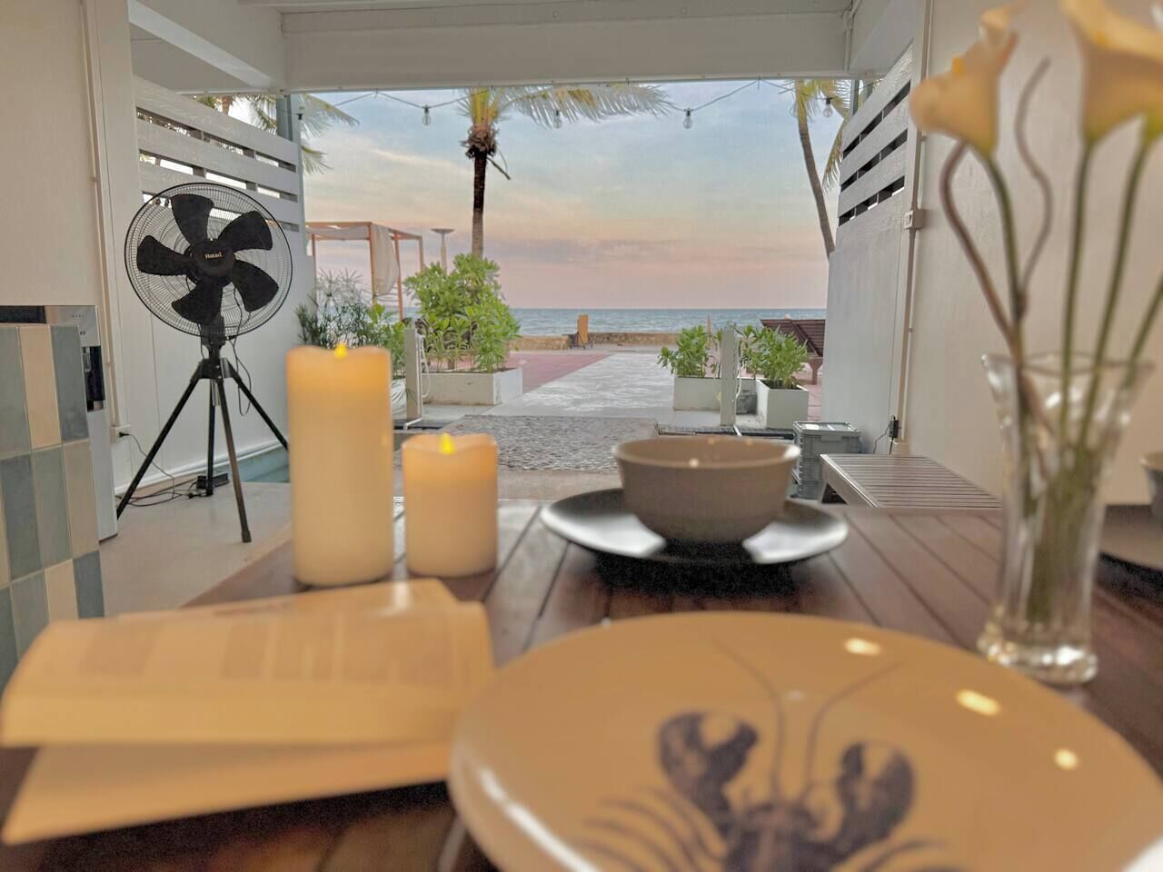 Uma área de refeições com vista para a praia, um ventilador, velas e um vaso com flores.