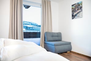 Ein Zimmer mit einem Bett, einem grauen Sofa, einem Fenster mit Vorhängen und einem Wandbild.