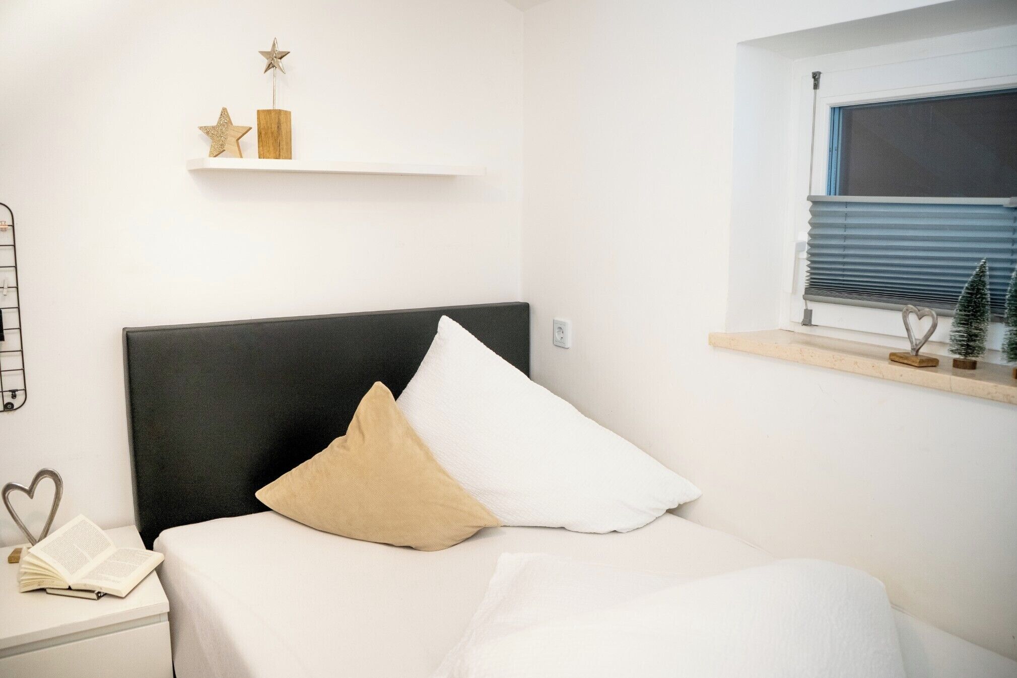 A bedroom with a bed, a nightstand with a book, a shelf with decorative items, and a window with blinds.