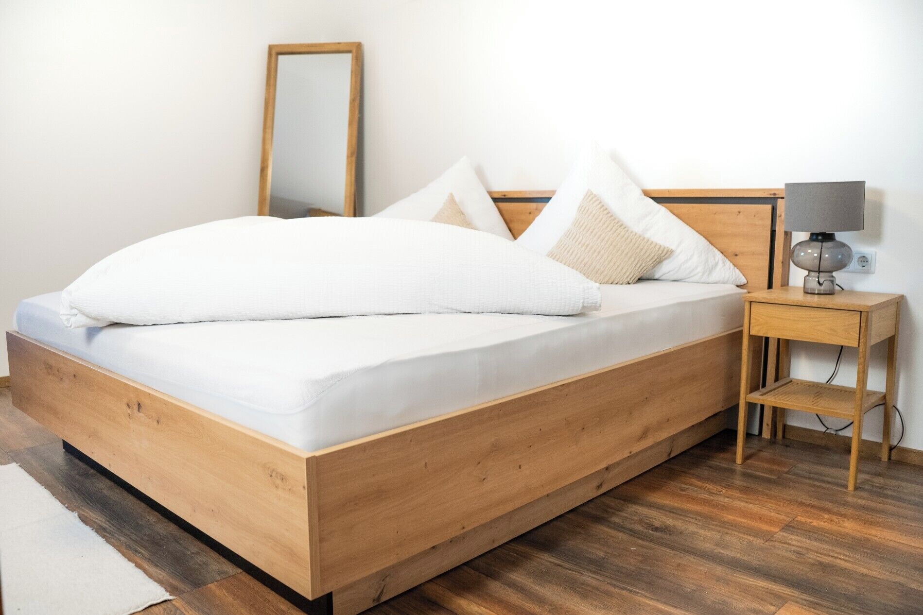 A wooden bed with white bedding, a bedside table with a lamp, and a mirror on the wall.