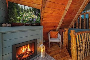 Ein gemütliches Blockhaus-Interieur mit Kamin, einem darüber hängenden Fernseher und einem Sessel am Fenster.