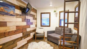 Ein Zimmer mit einer Holzwand, einem grauen Sofa, einem weißen, flauschigen Teppich und einem an der Wand befestigten Fernseher.