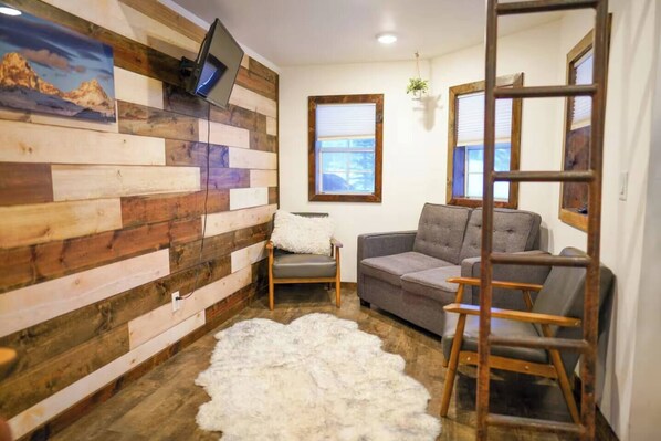 A room with a wooden wall, a gray sofa, a white furry rug, and a TV mounted on the wall.