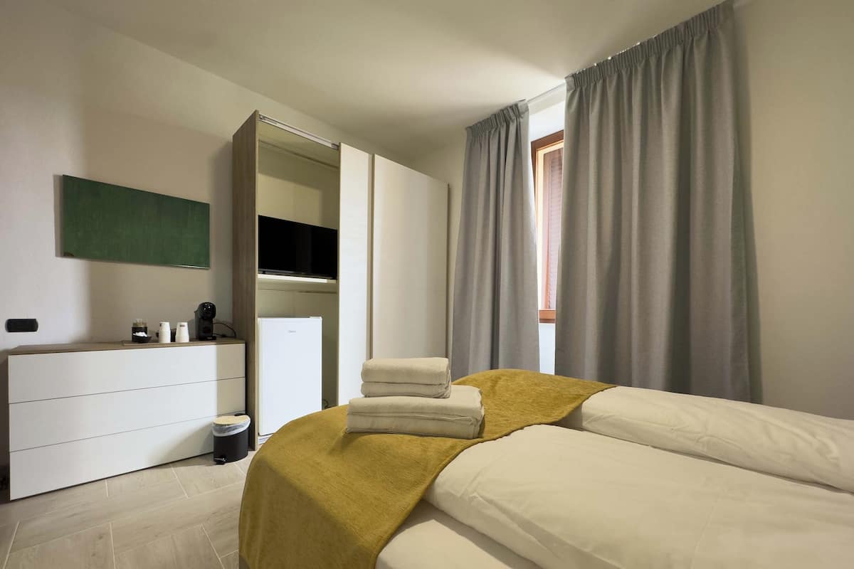A bedroom with a bed, a wardrobe, a mirror, a window with curtains, and a small table with items.