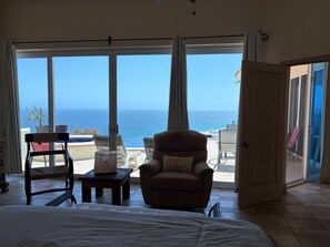 A bedroom with a large bed, a chair, a small table, and a view of the ocean through large windows. - Casa Amigos - Large 3 Bdrm Pedregal Beach Villa. (Cabo San Lucas)