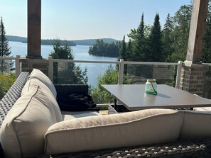 A balcony with a wicker sofa, a small table, and a view of a lake and forest.