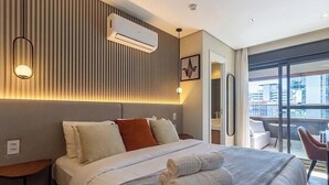 Quarto de hotel com uma cama, mesas de cabeceira, ar condicionado, luminária na parede e vista da cidade através de grandes janelas.