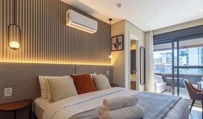 A hotel room with a bed, bedside tables, an air conditioning unit, a wall-mounted light, and a view of the city through large windows. - House w/ Garage in Rio Vermelho Tstedf0401 (Salvador)