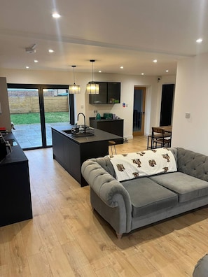 A modern kitchen with a central island, a grey sofa with a patterned throw, and a dining area with a wooden table and chairs. - Room-large Ensuite Double Room in Beauti (Chelmsford)