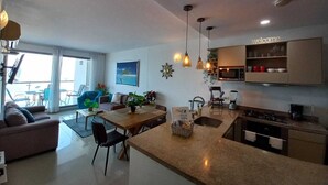 Eine moderne Küche mit einer zentralen Kochinsel, einem Essbereich mit Stühlen, einem Sofa und Blick ins Freie.