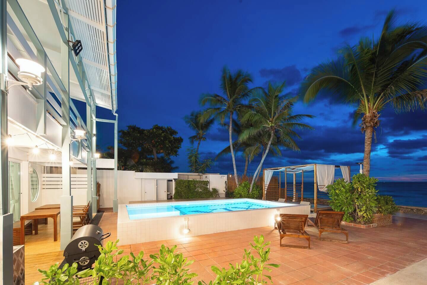 Zona de piscina exterior moderna con vista al océano y palmeras al anochecer.