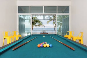 A pool table with balls, yellow chairs, and a view of the ocean through large windows.