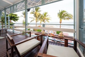 Uma área de estar coberta ao ar livre com mesas e bancos de madeira, com vista para uma praia com palmeiras e o oceano.