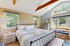 Ein Schlafzimmer mit Bett, Nachttischen, einem Schminktisch, einer Bank und großen Fenstern mit Blick auf Bäume.