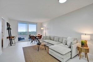 Ein modernes Wohnzimmer mit einem Ecksofa, einem Esstisch mit Stühlen, einer Stehlampe und Blick durch große Fenster.