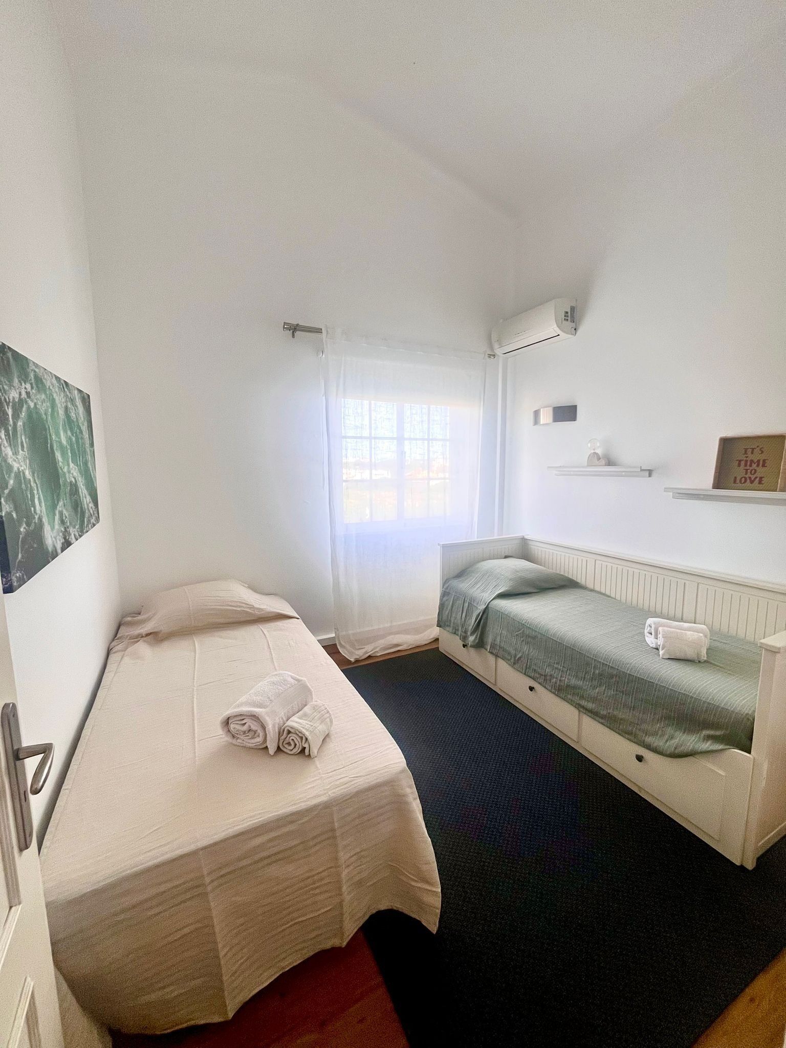A small, clean room with a single bed, a window with sheer curtains, a radiator, and a wall-mounted shelf with a sign.