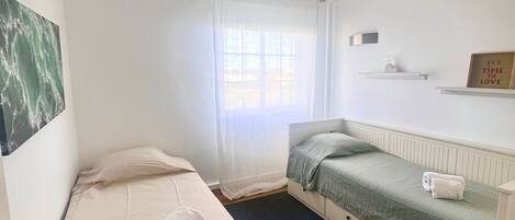 A small, clean room with a single bed, a window with sheer curtains, a radiator, and a wall-mounted shelf with a sign.