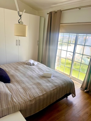 Ein Schlafzimmer mit einem großen Bett, einem Kleiderschrank, einem Fenster mit Vorhängen und Blick ins Freie.