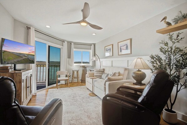 Ein modernes Wohnzimmer mit einer Couch, Sesseln, einem Fernseher, einem Deckenventilator und einem Balkon mit Blick.