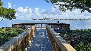 A wooden pier leading to a dock with a person fishing, overlooking a calm body of water and distant buildings. - Island Breeze Escape (Bradenton Beach)