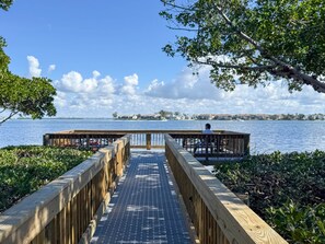 Eine hölzerne Pier führt zu einem Steg, wo eine Person fischt, mit Blick auf ruhiges Wasser und entfernte Gebäude.