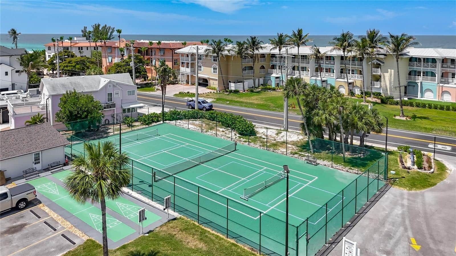 A tennis court surrounded by residential buildings, a parking area, and a road.