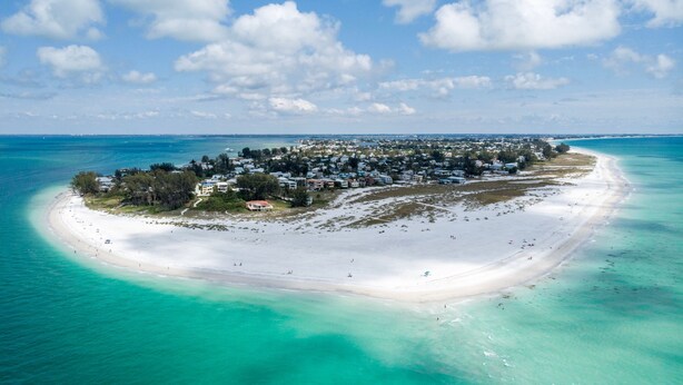 An aerial view of a coastal resort with a sandy beach, clear turquoise waters, and a cluster of buildings. - Island Breeze Escape (Bradenton Beach)