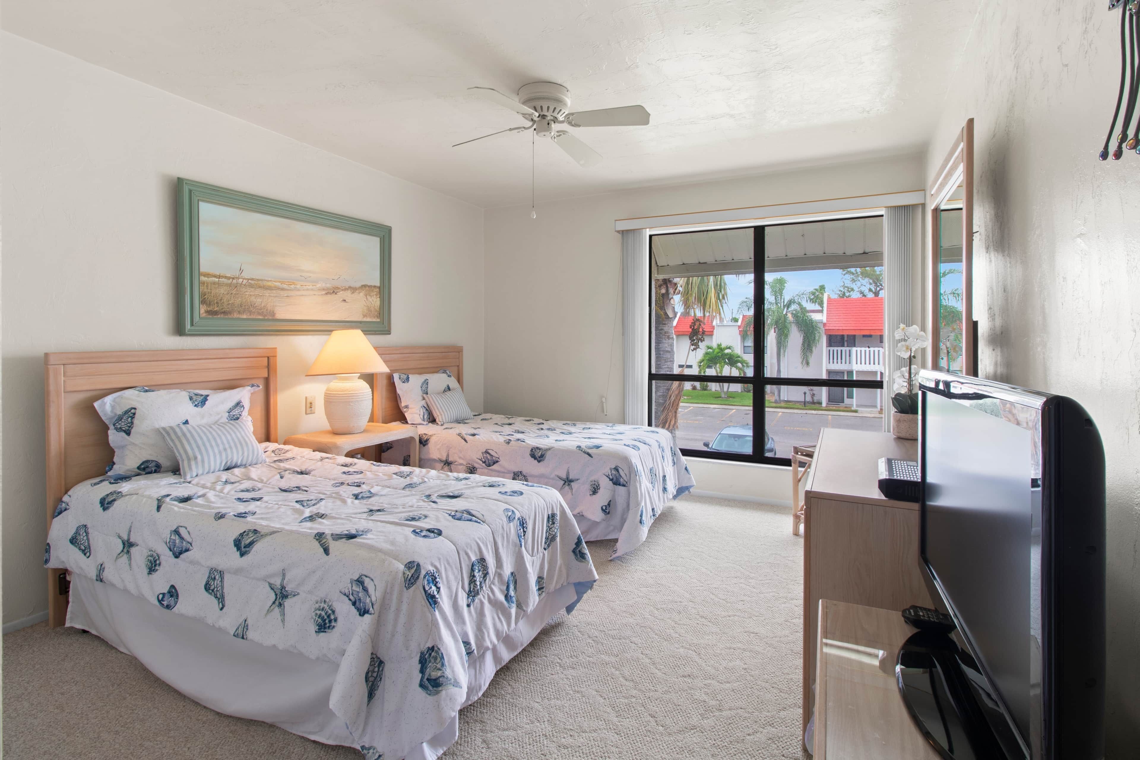 Habitación de hotel con dos camas, un escritorio con televisor, ventilador de techo y una ventana grande con vistas a palmeras.
