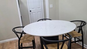 Une table de salle à manger ronde, entourée de quatre chaises, avec une porte visible au fond et un plancher en bois.