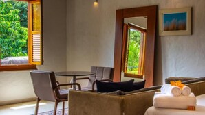 Une pièce comprenant un lit, une table et deux chaises. On y trouve des fenêtres avec des volets, un tableau accroché au mur et une vue sur la verdure à l’extérieur.
