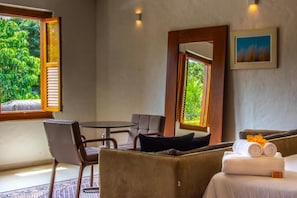 A room with a bed, a table, and two chairs. There are windows with shutters, a painting on the wall, and a view of greenery outside.