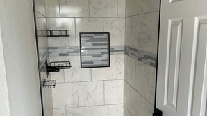 Una ducha a nivel con puerta de vidrio, azulejos de mármol blanco, un cabezal de ducha y un cuadro en la pared.