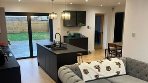 A modern kitchen with a central island, a grey sofa with a patterned throw, and a dining area with a wooden table and chairs. - Room-bright Double Room in Chelmsford Ho (Chelmsford)