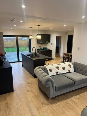 A modern kitchen with a central island, a grey sofa with a patterned throw, and a dining area with a wooden table and chairs. - Room-bright Double Room in Chelmsford Ho (Chelmsford)