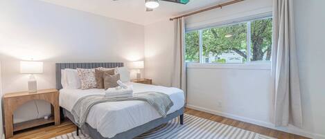 A bedroom with a bed, bedside tables, a ceiling fan, a window with curtains, and a view of trees outside.