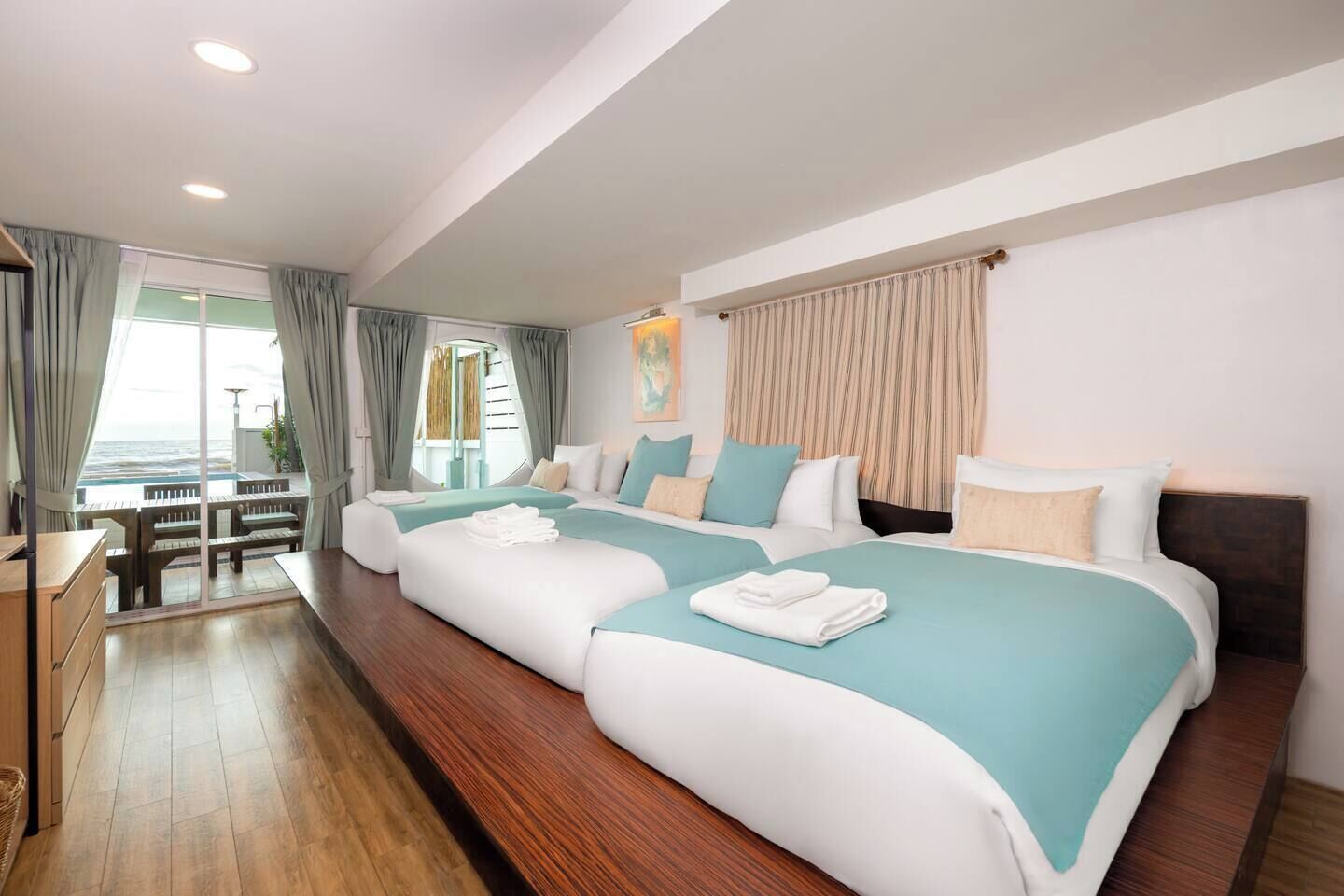 A hotel room with two beds, a wooden floor, a view of the sea, and a balcony with a table and chairs.