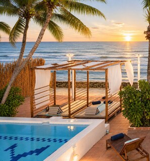 Área junto a la piscina con una pérgola de madera, sillas de descanso y vista al océano al atardecer.