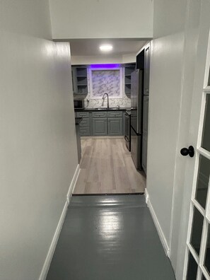 Un couloir étroit mène à une cuisine dotée de meubles gris, d’un évier et d’un réfrigérateur.