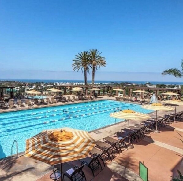 A swimming pool with sun loungers and umbrellas, surrounded by palm trees and a clear view of the ocean.