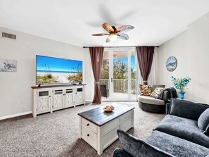 A living room with a TV displaying a beach scene, a ceiling fan, a sofa, a coffee table, and a sliding glass door leading to an outdoor area. - Family Resort, Garden View 3rd FL, Pools, Beach walk and shuttle (Destin)