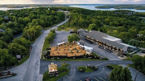 An aerial view of a property with a parking lot, a building with a flat roof, and a surrounding forested area. - Suites at Waterside (Branson)